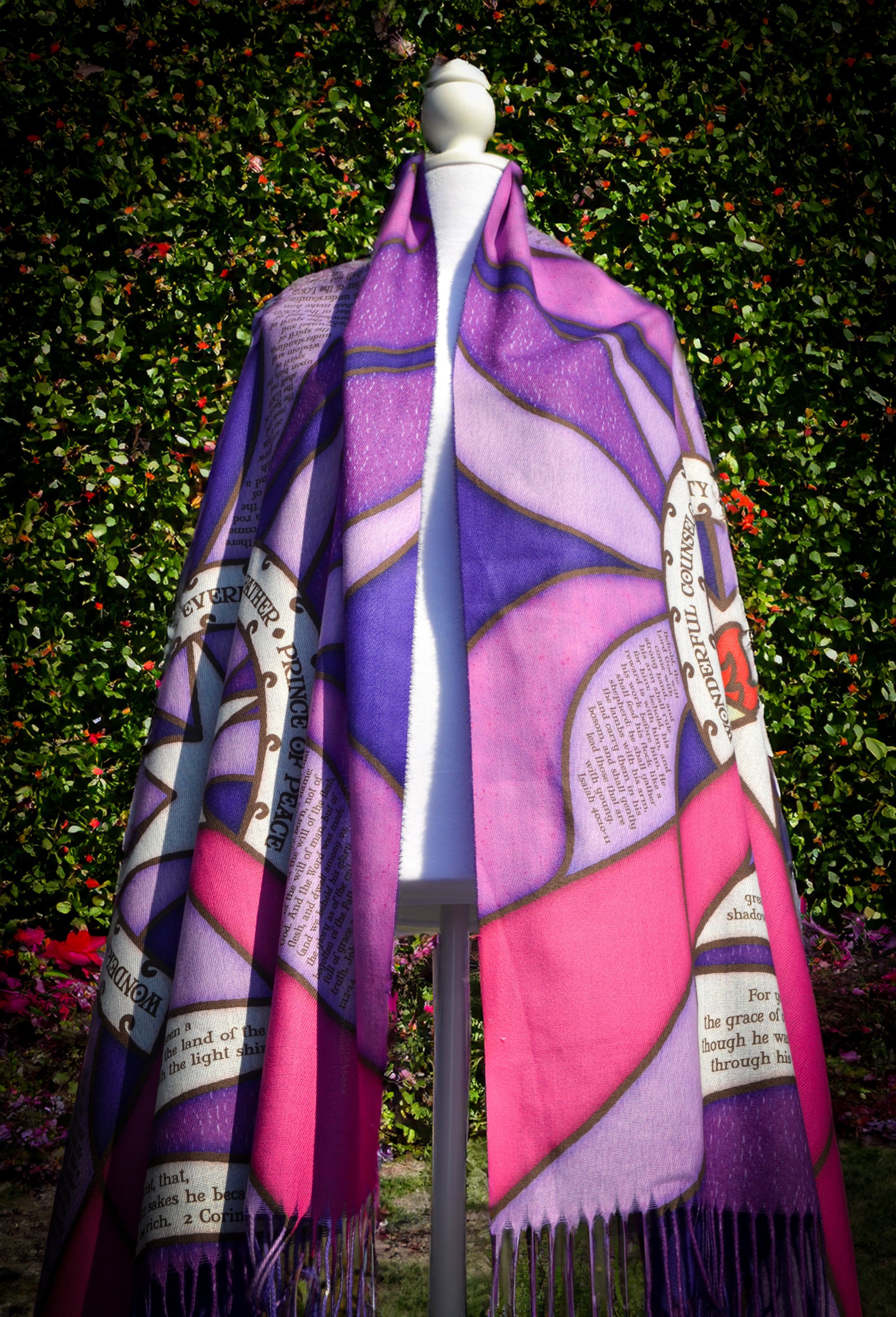 Purple Advent Prayer Scarf by Prayer Scarves® shown draped on a mannequin outdoors, highlighting stained-glass–style purple and pink sections, black lead-style lines, a central flame design, and printed scripture elements.