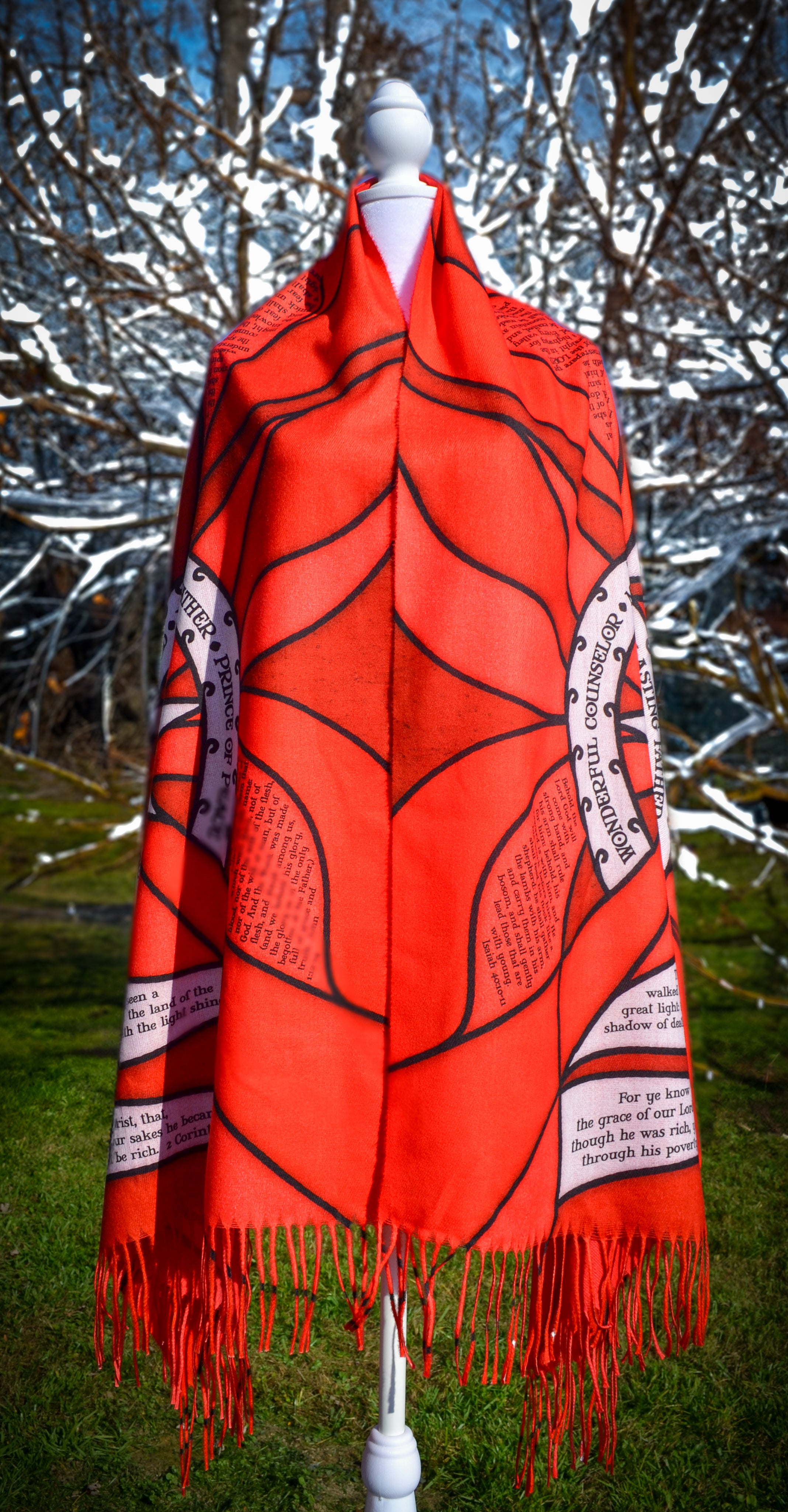 Red Advent Prayer Scarf by Prayer Scarves® displayed on a mannequin outdoors, showing bold red stained-glass–style panels, black lead-style outlines, and printed scripture text along the design.