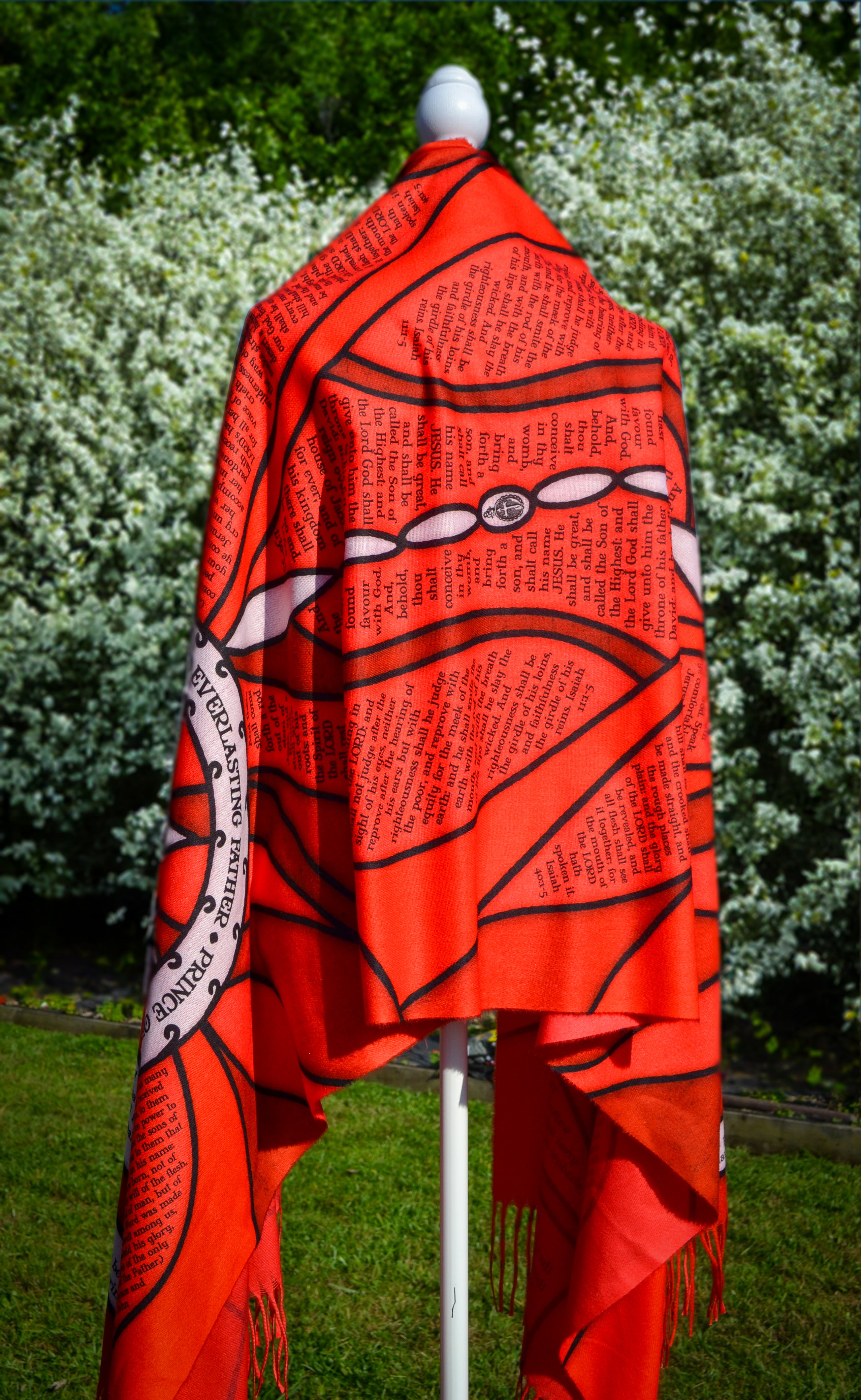 Red Advent Prayer Scarf by Prayer Scarves® displayed on a mannequin outdoors, showing bold red stained-glass–style panels, black lead-style outlines, and printed scripture text along the design.