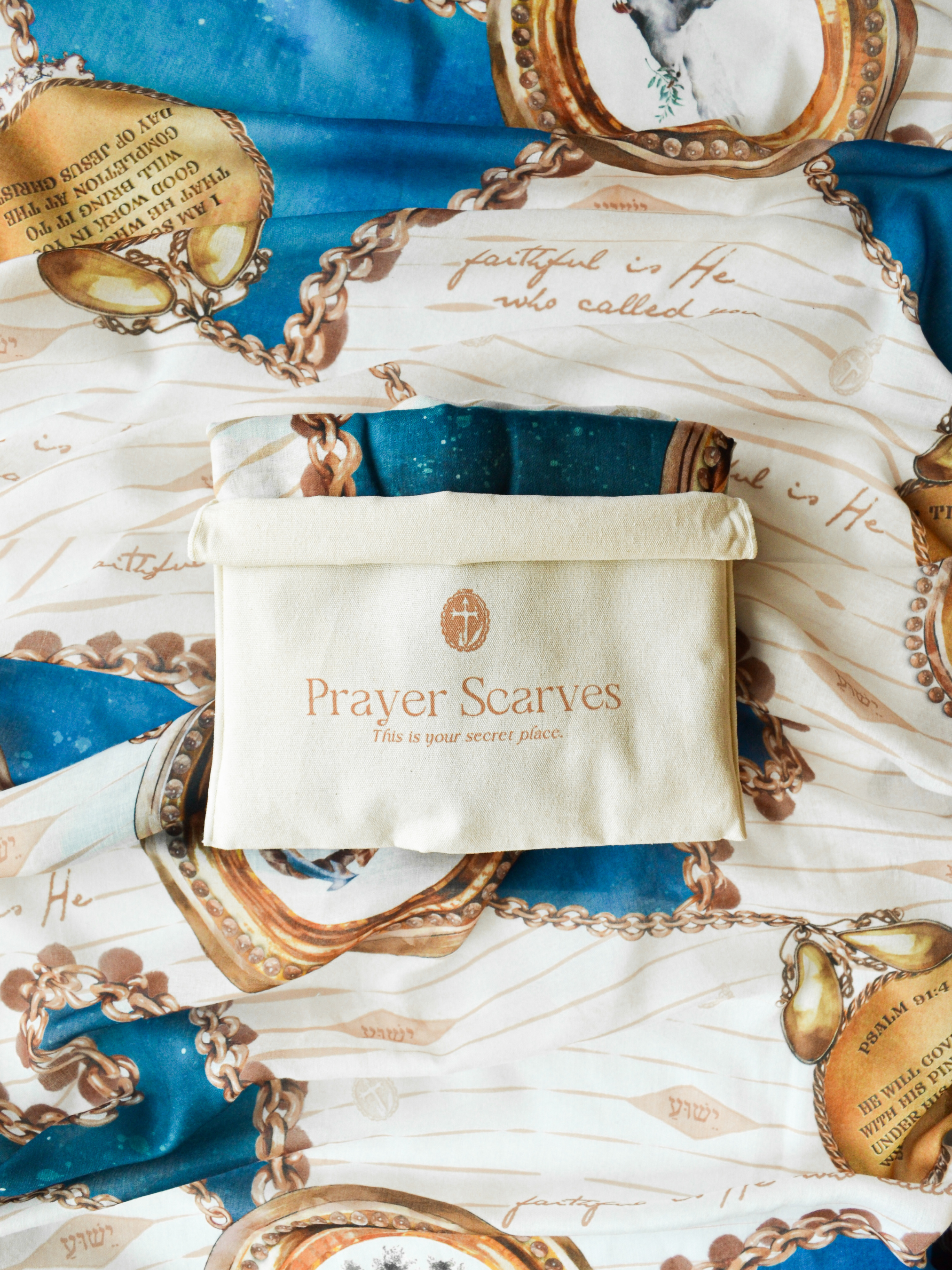 Folded Faithfulness Prayer Scarf by Prayer Scarves® shown beneath a beige fabric storage pouch printed with the Prayer Scarves logo, featuring blue and teal sections, gold chain motifs, circular medallion designs, handwritten-style script details, and visible printed scripture text.
