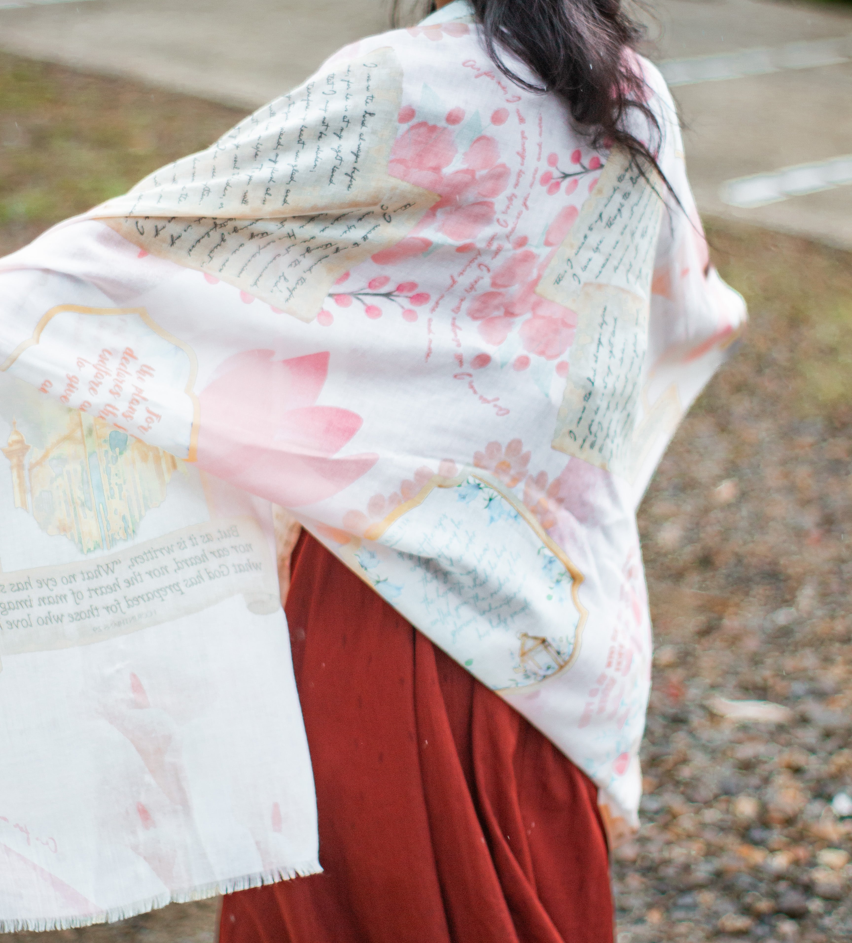 Hope prayer scarf by Prayer Scarves® worn outdoors, shown draped over the head and shoulders, featuring soft pink and cream tones, handwritten-style scripture text, gentle color washes, and illustrated design elements visible on the fabric.