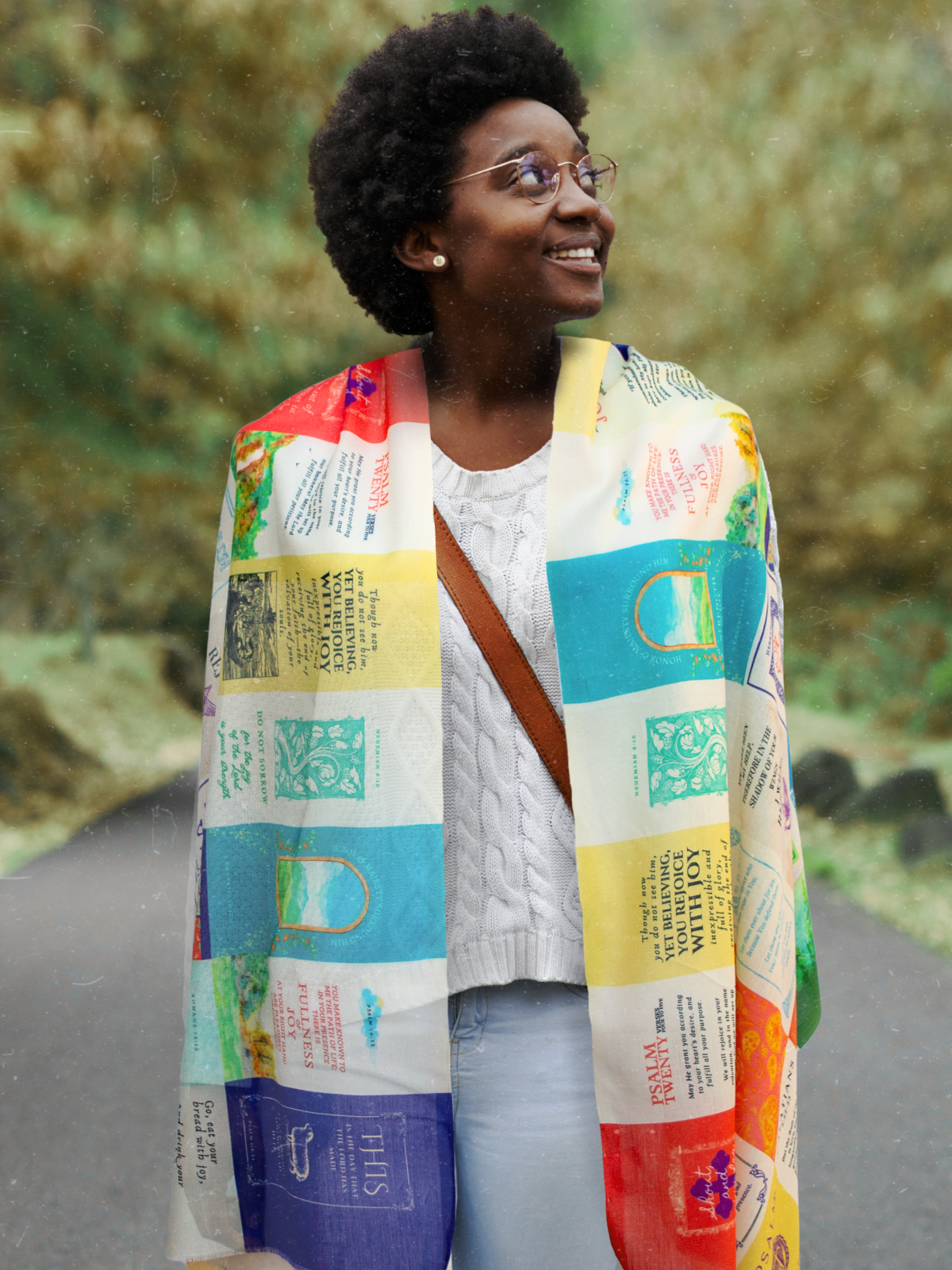 Joy Cotton prayer scarf by Prayer Scarves® worn outdoors by a model, featuring a multicolor patchwork design with scripture text blocks, decorative motifs, and varied patterned panels draped over the shoulders.