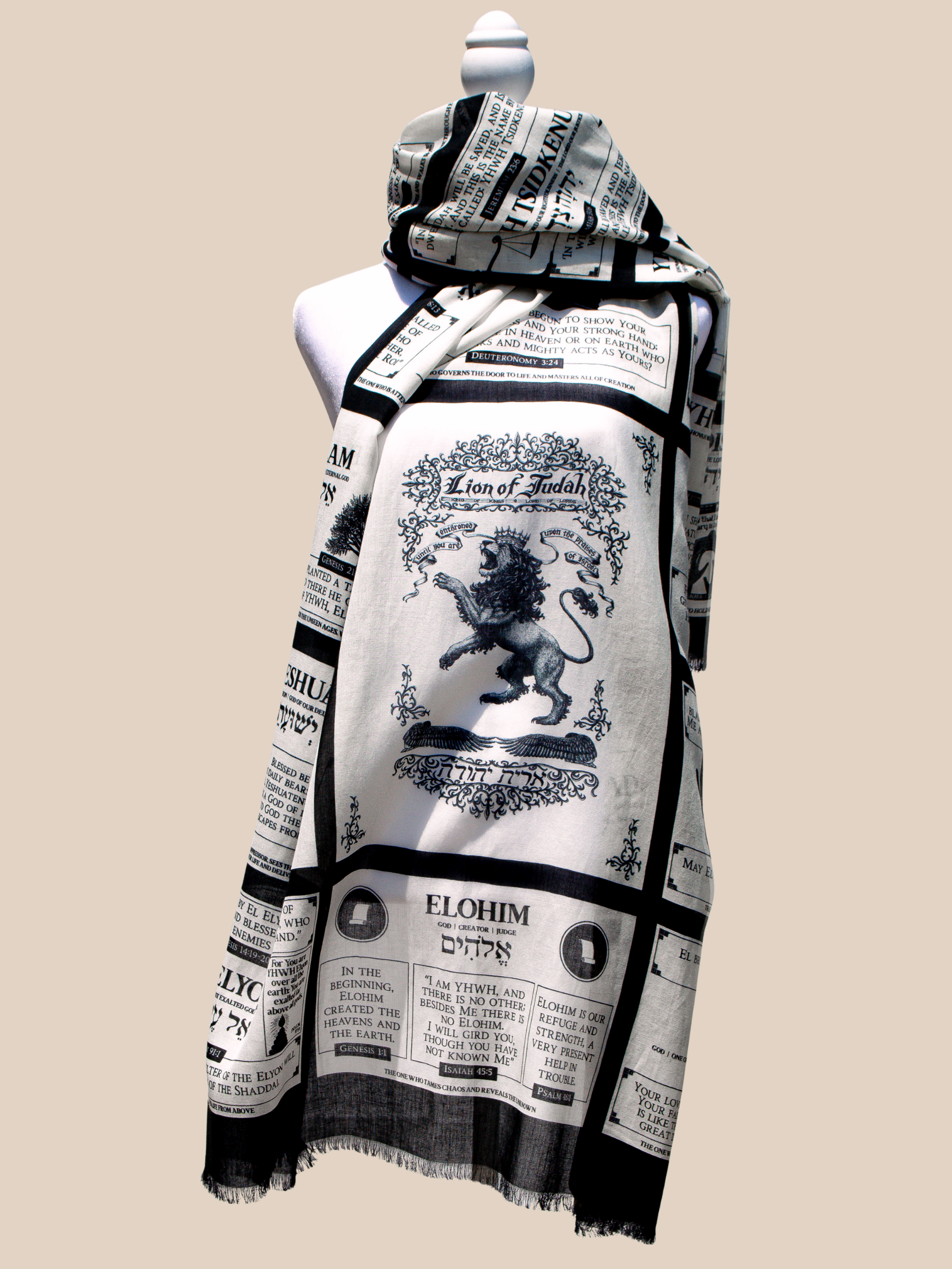 A white and black Names of God protection prayer scarf styled on a mannequin, prominently featuring a detailed "Lion of Judah" illustration. The fabric of the Prayer Scarves® includes a grid of Hebrew names for God, such as Elohim, alongside Scripture passages about God’s protection, strength, and role as Creator.
