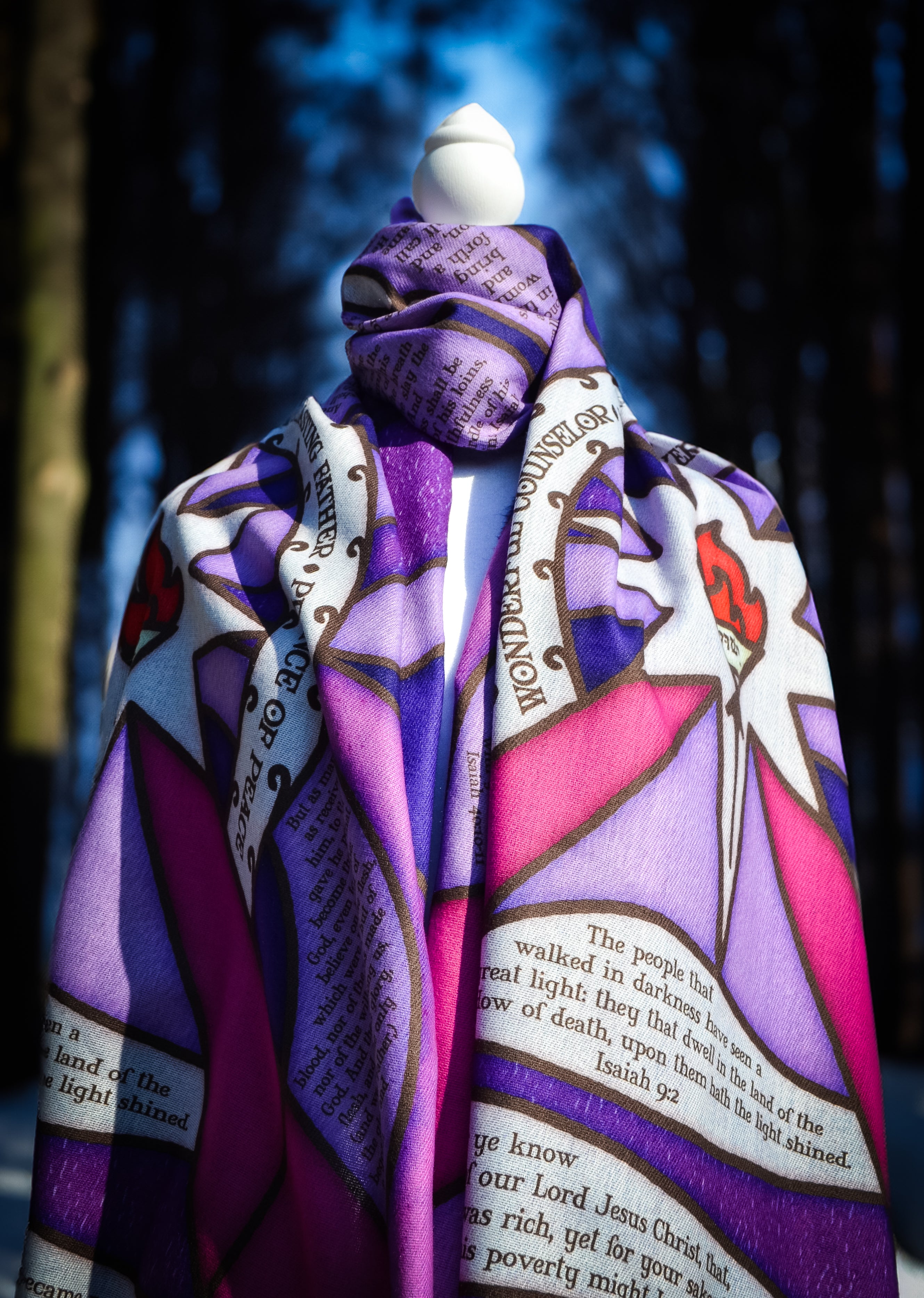 Purple Advent Prayer Scarf by Prayer Scarves® shown draped on a mannequin outdoors, highlighting stained-glass–style purple and pink sections, black lead-style lines, a central flame design, and printed scripture elements.