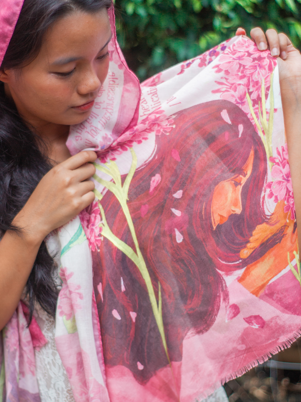 Adoration prayer scarf by Prayer Scarves® shown worn outdoors, featuring an illustrated depiction of Mary, pink floral blossoms, green botanical stems, soft pink and cream tones, and visible scripture text across the fabric.