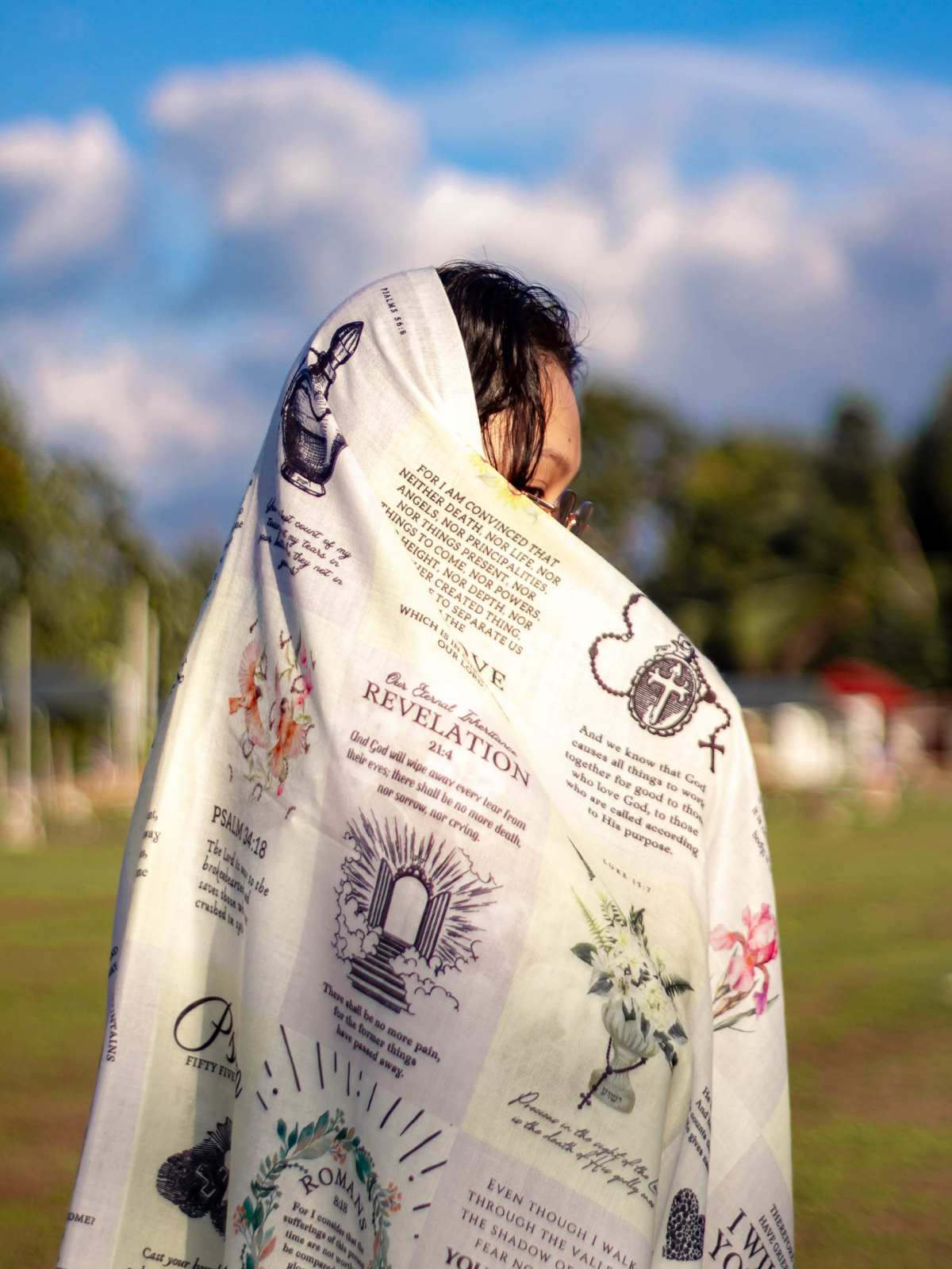 Dawn prayer scarf by Prayer Scarves® worn outdoors and draped around a person’s shoulders, featuring an ivory background with printed scripture passages, botanical illustrations, and visible text including “I am with you,” Psalm references, Romans, and 2 Corinthians.