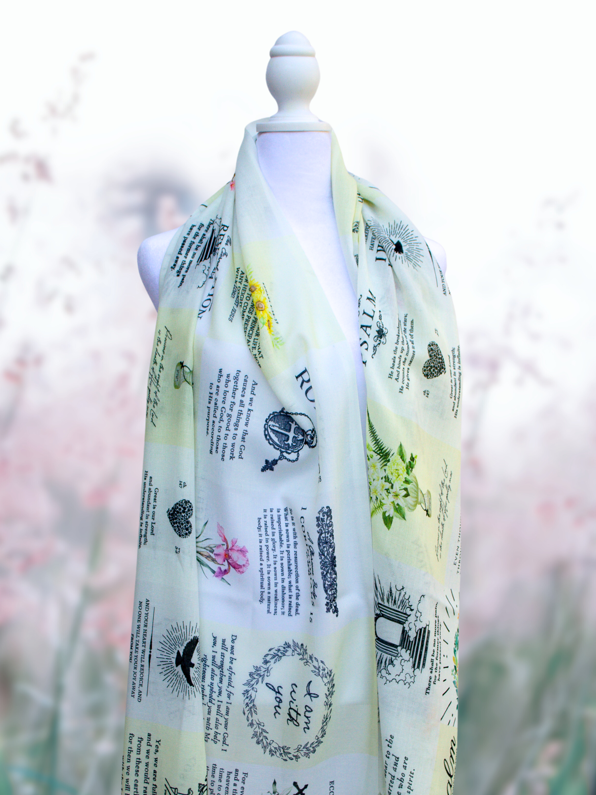 Dawn prayer scarf by Prayer Scarves® displayed draped symmetrically over a white mannequin, featuring a soft ivory and pale yellow design with scattered scripture excerpts, botanical illustrations, and visible text elements including Romans, Psalm references, and the phrase “I am with you.”