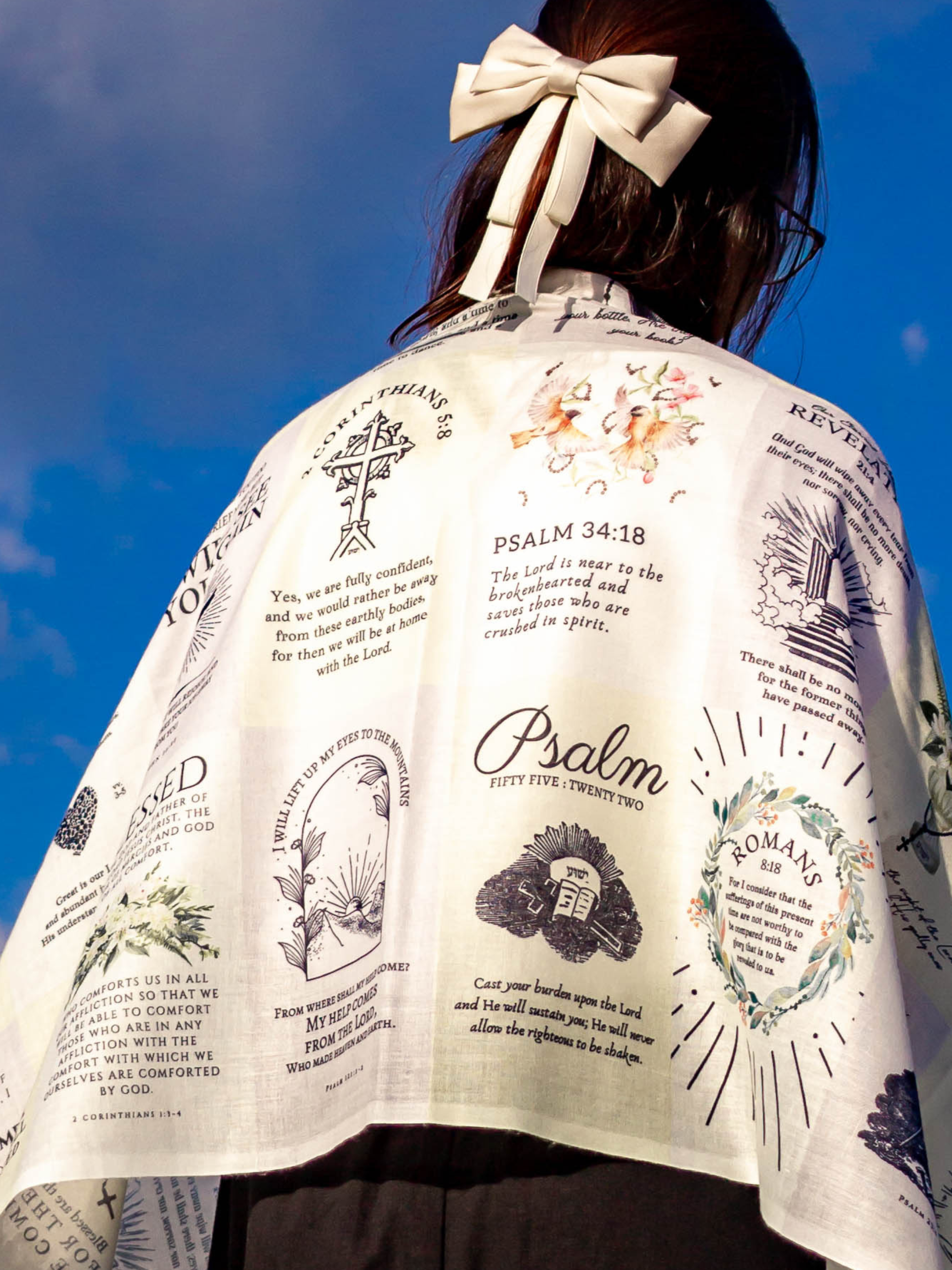 Dawn prayer scarf by Prayer Scarves® worn outdoors and draped around a person’s shoulders, showing an ivory-toned design with printed scripture excerpts, botanical illustrations, and visible text elements including “I am with you,” Psalm references, Romans, and 2 Corinthians.