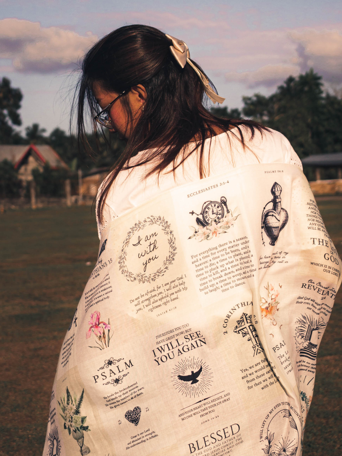 Dawn prayer scarf by Prayer Scarves® worn outdoors and draped around a person’s shoulders, featuring an ivory background with printed scripture passages, botanical illustrations, and visible text including “I am with you,” Psalm references, Romans, and 2 Corinthians.