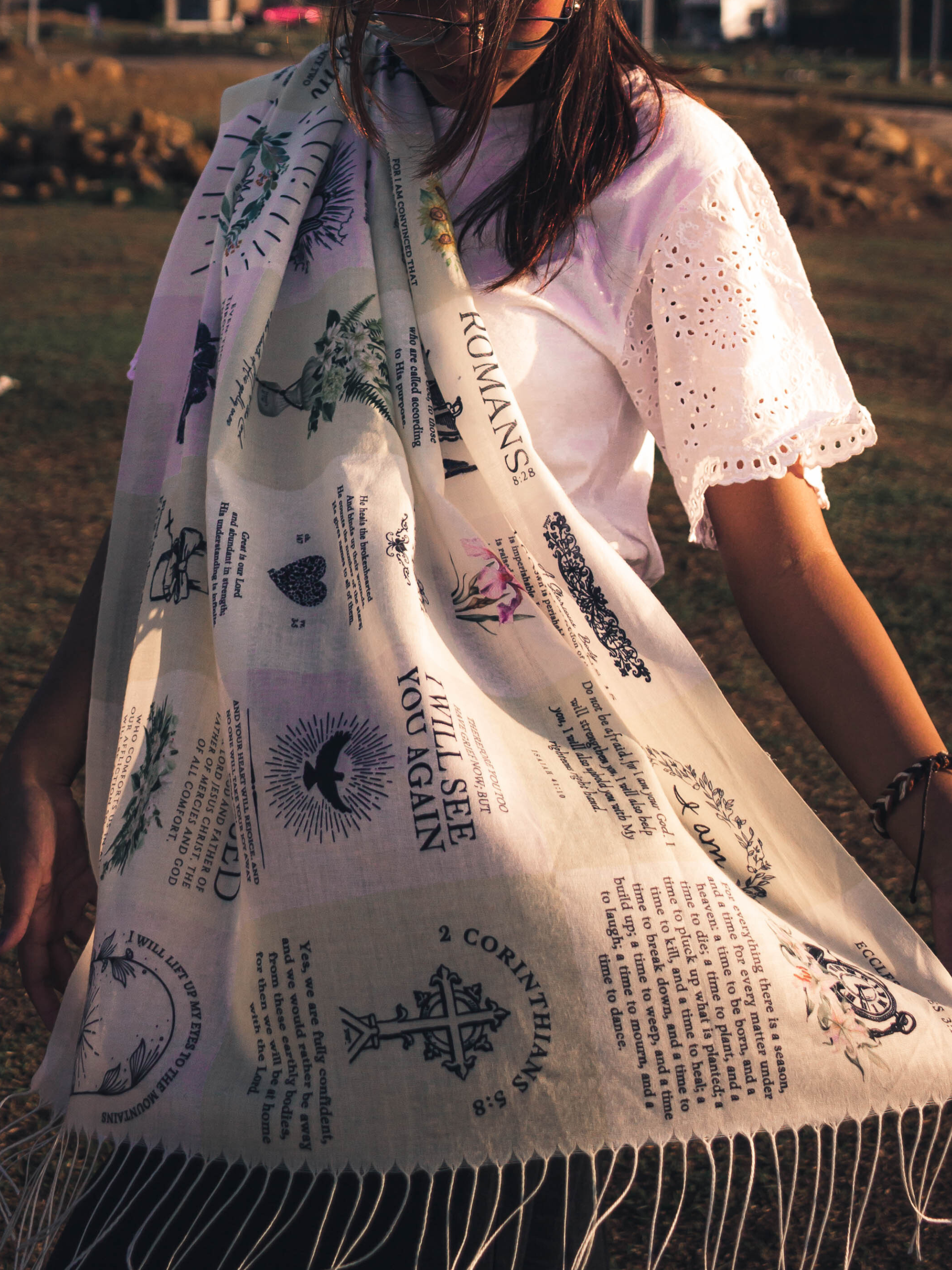 Dawn prayer scarf by Prayer Scarves® worn outdoors and draped around a person’s shoulders, showing an ivory-toned design with printed scripture excerpts, botanical illustrations, and visible text elements including “I am with you,” Psalm references, Romans, and 2 Corinthians.
