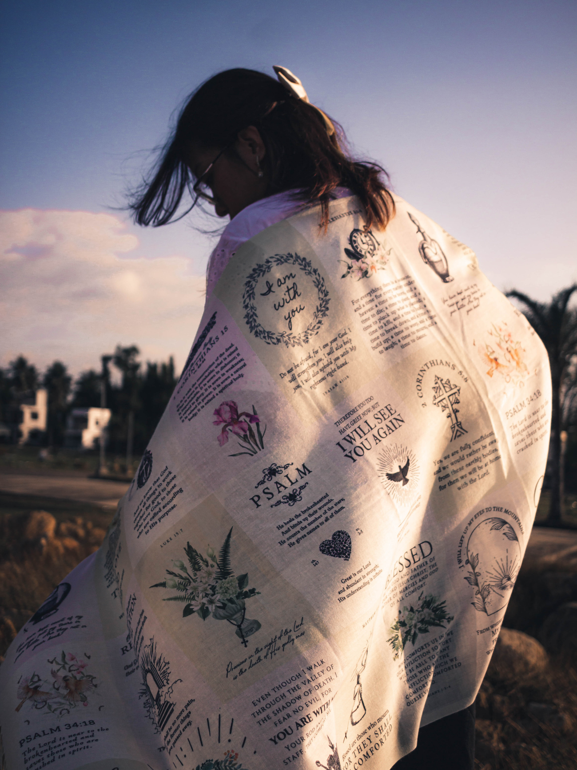 Dawn prayer scarf by Prayer Scarves® worn outdoors and draped around a person’s shoulders, showing an ivory-toned design with printed scripture excerpts, botanical illustrations, and visible text elements including “I am with you,” Psalm references, Romans, and 2 Corinthians.
