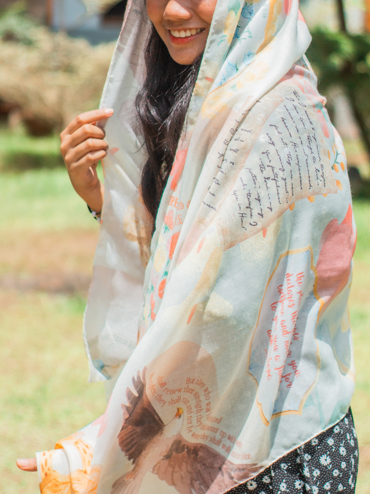 Hope prayer scarf by Prayer Scarves® worn outdoors in the honeysuckle colorway, featuring soft peach, yellow, and blue floral elements, framed scripture text panels, handwritten-style typography, and illustrated motifs visible across the fabric.