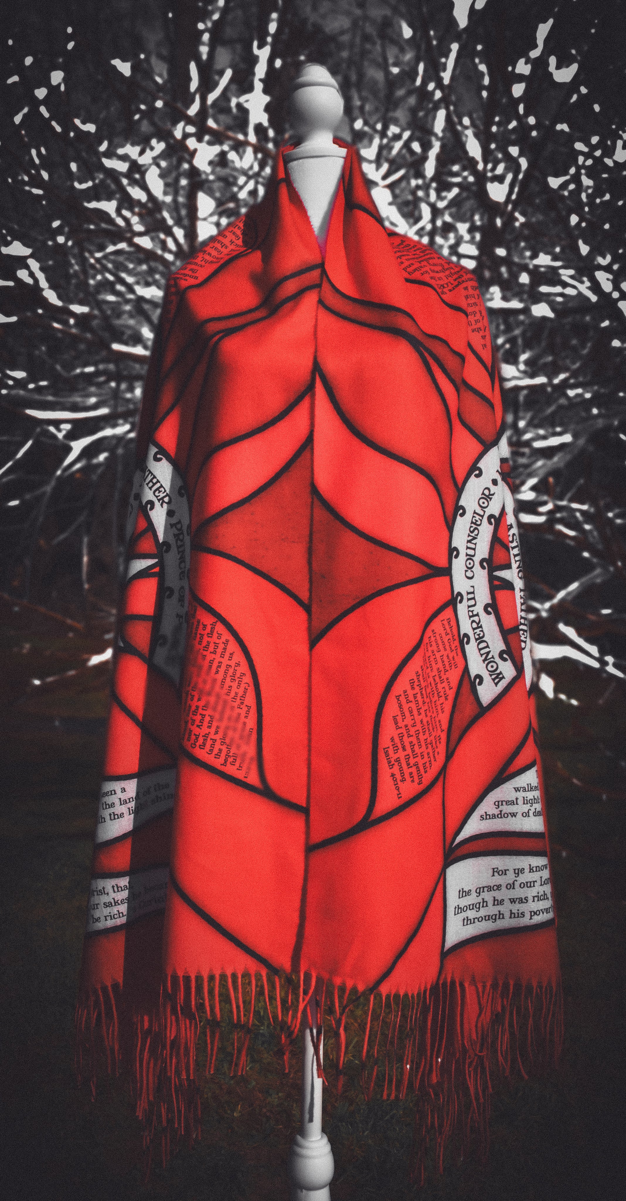 Red Advent Prayer Scarf by Prayer Scarves® displayed on a mannequin outdoors, showing bold red stained-glass–style panels, black lead-style outlines, and printed scripture text along the design.