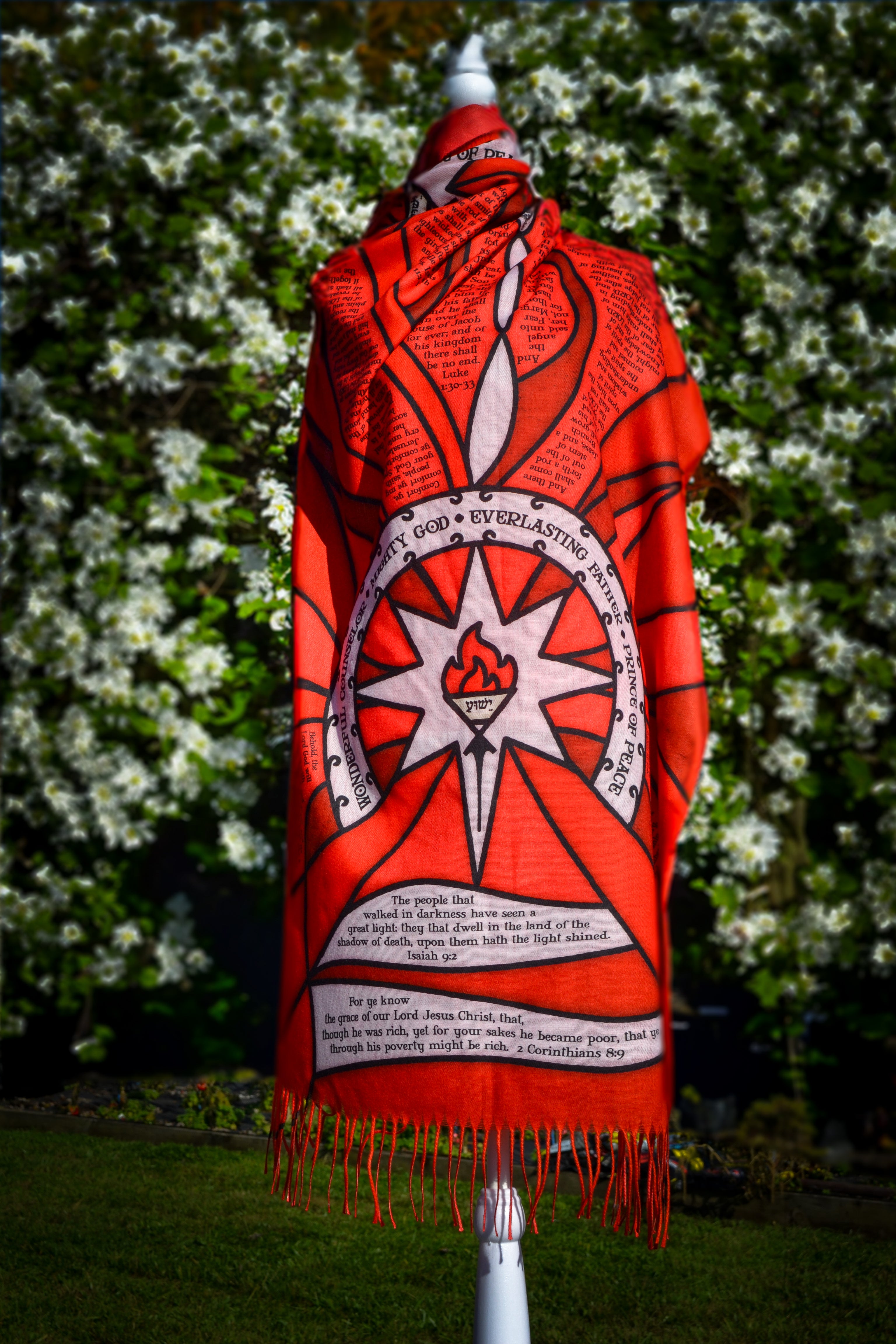 Red Advent Prayer Scarf by Prayer Scarves® displayed on a mannequin outdoors, showing bold red stained-glass–style panels, black lead-style outlines, and printed scripture text along the design.