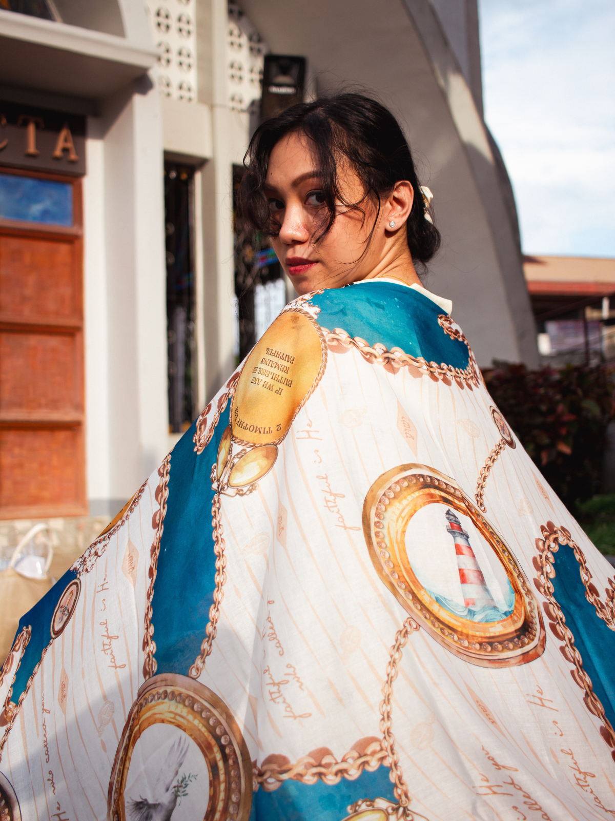 Person wearing the Faithfulness Prayer Scarf by Prayer Scarves® draped over the shoulders outdoors, featuring blue and teal sections, gold chain detailing, circular medallion imagery, and visible scripture text.