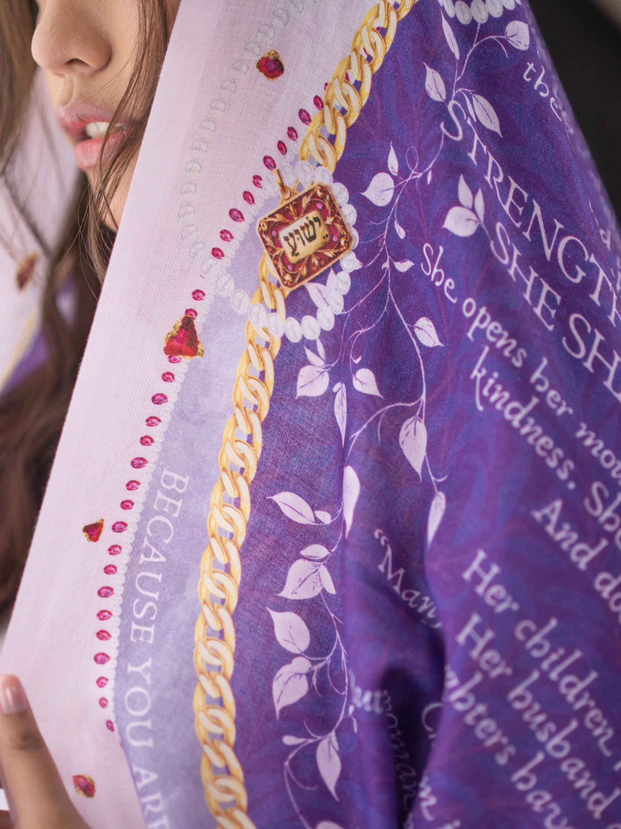 Prayer Scarves® Proverbs 31 Virtuous Woman purple scarf detail showing scripture text, white floral vine illustrations, gold-toned chain-style border accents, small jewel-like motifs, and a decorative pendant element with Hebrew lettering along the edge.