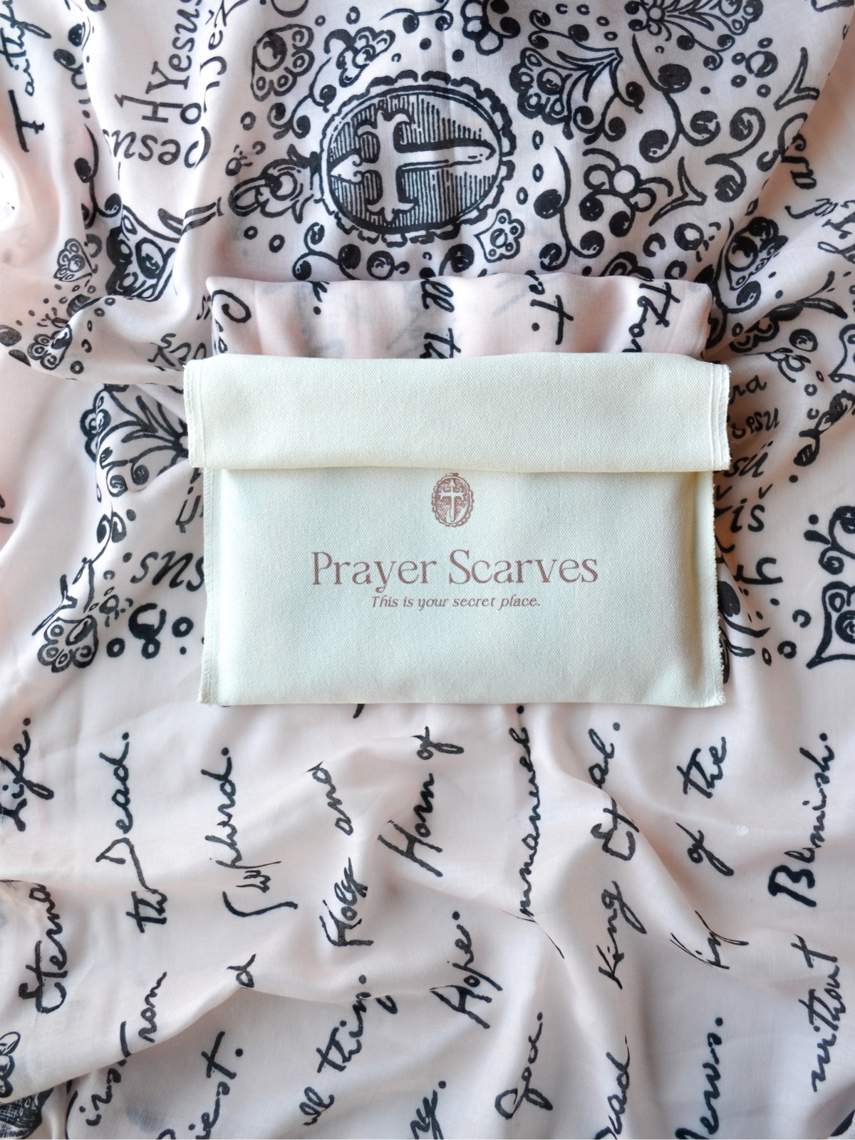 The And He Shall Be Called prayer scarf by Prayer Scarves®, shown with its included fabric storage pouch placed on the scarf, highlighting the detailed names and titles of Jesus throughout the design