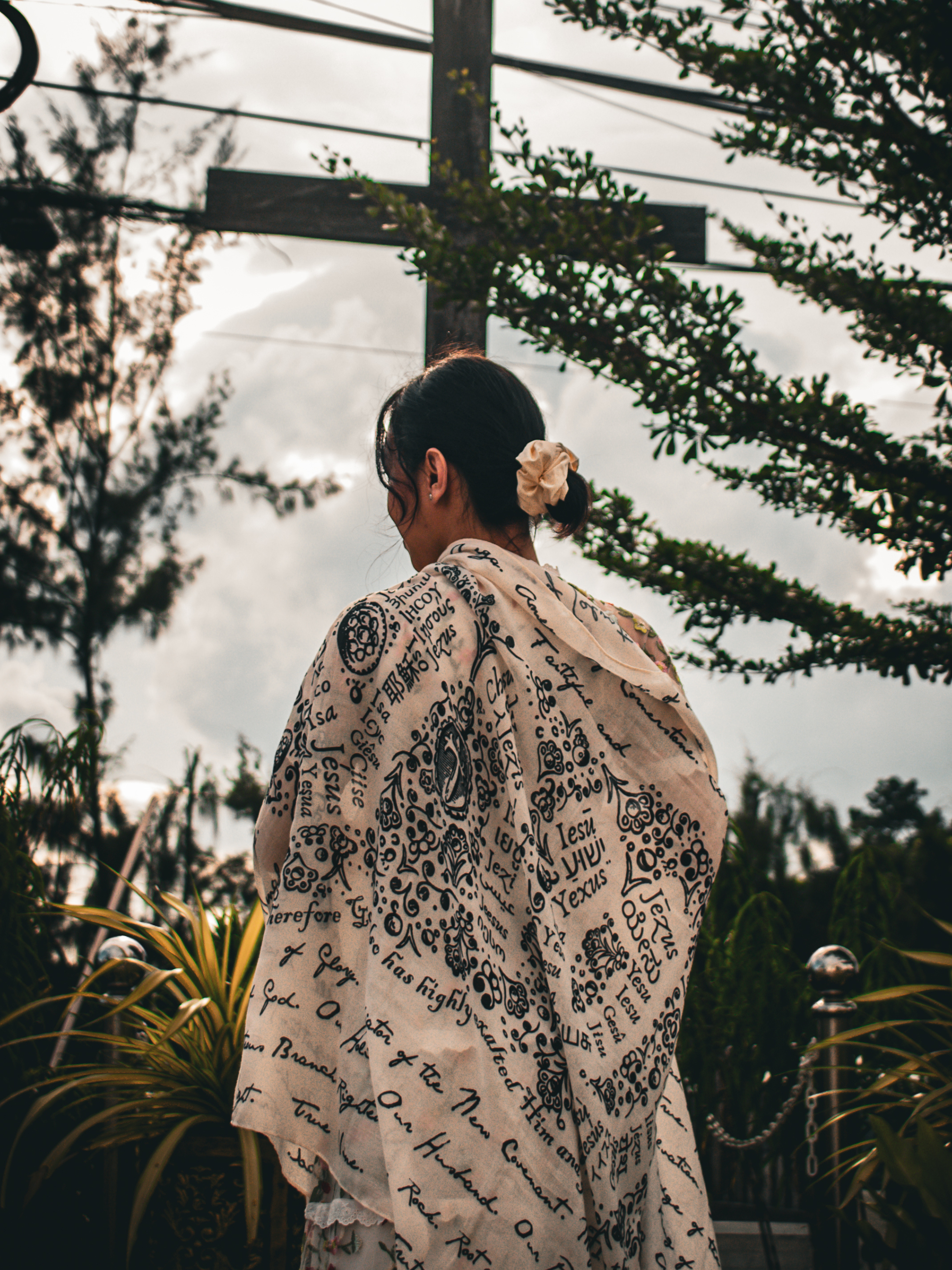 And He Shall Be Called prayer scarf by Prayer Scarves® wrapped around a woman standing outdoors near a cross, featuring handwritten and illustrated names and titles of Jesus throughout the design