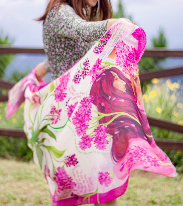 Adoration prayer scarf by Prayer Scarves® shown worn outdoors as a model holds the scarf open, featuring an illustrated depiction of Mary, bright pink floral clusters, green botanical stems, cream background tones, and visible scripture text throughout the design.